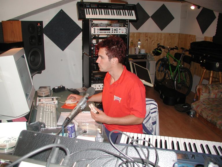 Ein Bandmitglied arbeitet am Studiocomputer; Mikrofon, Keyboard und Studiotechnik stehen rundherum.