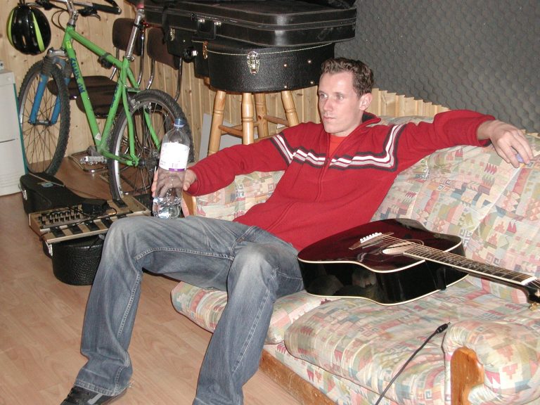 Ein Bandmitglied sitzt in einer Studiopause mit Akustikgitarre auf dem Sofa.