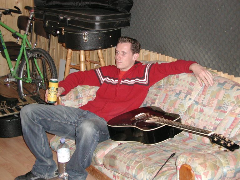 Ein Bandmitglied sitzt mit Akustikgitarre auf dem Sofa im Regieraum.
